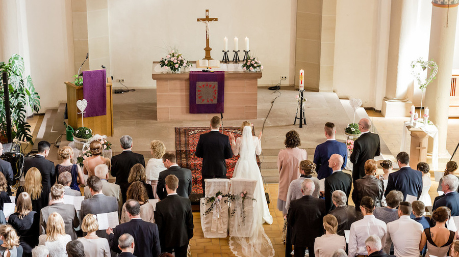 Zu sehen ist ein voller Kirchsaal, festlich eingerichtet für eine Hochzeit. Die anwesenden Gäste und das Brautpaar haben sich erhoben, um gemeinsam zu beten/ singen.